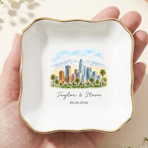 Puede incluir: Un plato cuadrado de cerámica blanca con un borde festoneado dorado. El centro presenta una ilustración en acuarela de un horizonte urbano con palmeras y montañas, con el texto Taylor & Steve y la fecha 06.06.2026 debajo.