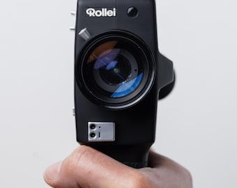 May include: A black Rollei Super 8 movie camera with a large lens and silver accents is held in a hand against a white background. The camera lens reflects blue and orange light.