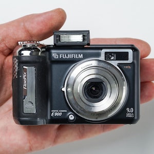 May include: A black Fujifilm E900 digital camera with a silver lens and flash. The camera has the text "FUJIFILM" and "FinePix" on the body. It is a 9.0 megapixel camera.