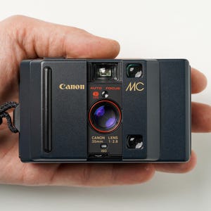 May include: A black Canon MC 35mm film camera with gold lettering, held in a hand. The camera has a lens with a red ring and the words "AUTO FOCUS" above it. The camera strap is black.