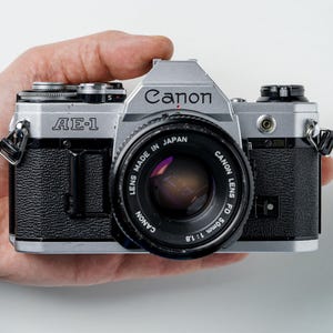 Puede incluir: Una cámara de película Canon AE-1 plateada y negra con una lente de 50 mm. La cámara se sostiene en una mano y presenta el logotipo de Canon y el nombre del modelo "AE-1". La lente está marcada "LENS MADE IN JAPAN CANON LENS FD 50mm 1:1.8".