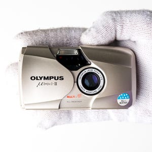 May include: A champagne-coloured Olympus mju: II 35mm film camera with a sliding lens cover. The front features the text OLYMPUS mju: II, MULTI AF, ALL-WEATHER, and a circular sticker with a blue raindrop icon and the text ALL-WEATHER.
