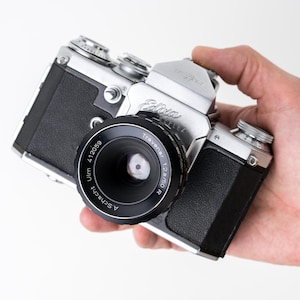 May include: A vintage Edixa Reflex 35mm film camera with a silver metal body and black textured leatherette grip. The camera is equipped with a black A. Schacht Ulm Travenar 1:2.8/50 R lens featuring visible aperture blades and white text.