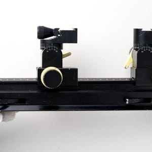 May include: A black metal macro focusing rail with two adjustable sliding carriages. The rail features a white printed scale along the top edge, two black and cream-colored adjustment knobs, and two locking levers on each sliding assembly.
