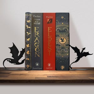 May include: Four books with the titles "Eragon" and "Eldest" are held in place by two black metal dragon-shaped bookends. The books have gold and red spines. The bookends are on a wooden shelf.