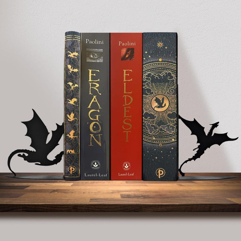 May include: Four books with the titles "Eragon" and "Eldest" are held in place by two black metal dragon-shaped bookends. The books have gold and red spines. The bookends are on a wooden shelf.
