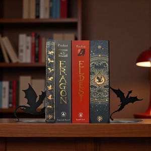 May include: A collection of fantasy books, including "Eragon" and "Eldest", held between two black metal dragon-shaped bookends. The books have gold lettering and decorative covers, set on a wooden surface.