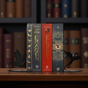 May include: A collection of books, including "Eragon" and "Eldest", standing upright on a wooden shelf. Black metal dragon-shaped bookends support the books. The books have gold and red spines.