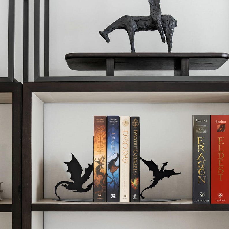May include: A bookshelf with a dark wooden frame and white interior. The shelf displays several books held by black metal dragon-shaped bookends. A dark sculpture of a horse and rider sits on the top shelf.