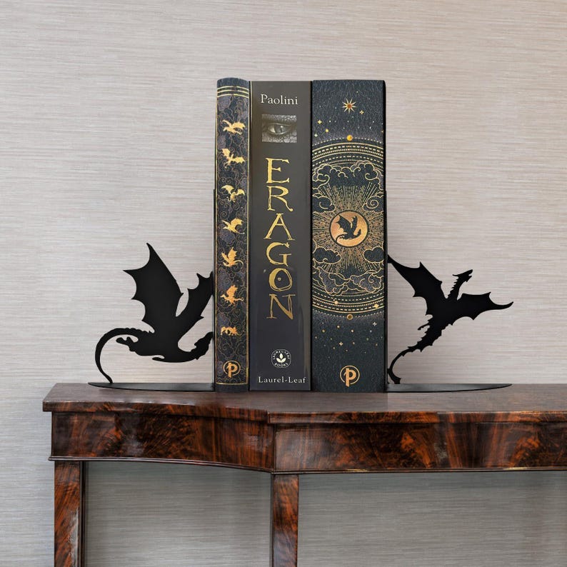 May include: Black metal dragon bookends flanking three books on a wooden table. The books are dark with gold lettering and accents. The bookends are in the shape of dragons with outstretched wings.