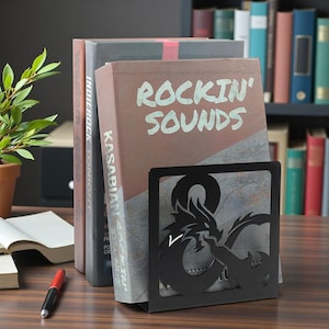 Peut inclure: Un serre-livres en métal noir avec un motif de dragon, tenant trois livres. Les livres sont intitulés "Rockin' Sounds" et "Kasabian". Un stylo et une petite plante en pot sont également visibles.