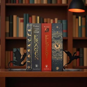 May include: Four hardcover books with the titles "Eragon" and "Eldest" are displayed between two black metal dragon-shaped bookends. The books are on a wooden shelf in front of a bookshelf.