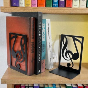 May include: Black metal bookends with a musical note design. The bookends are holding several books with titles including "Western Rodeo" and "The Art of Kinfolk". The books are on a wooden shelf.