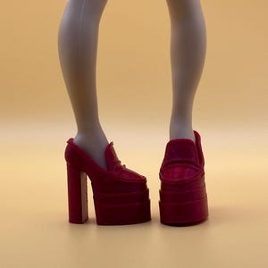May include: A pair of red platform heels with a block heel and a loafer-style design. The shoes are on a doll's legs, with a neutral background.