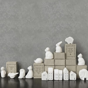 Puede incluir: Una colección de figuritas de cerámica blanca, que incluyen animales y casas en miniatura, dispuestas sobre una superficie oscura contra un fondo gris. Algunas figuritas están colocadas sobre pequeñas cajas con los textos "Flipping love you", "Sometimes all you need is a big bear hug" y "Love ave to the moon and back".