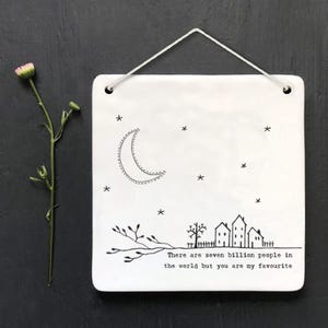 以下が含まれることがあります： 白のセラミック製の正方形の壁掛け。黒と白で三日月、星、家が描かれています。テキストには「世界には70億人の人々がいますが、あなたは私のお気に入りです」と書かれています。