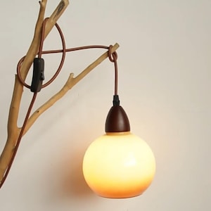 Puede incluir: Una lámpara artesanal única con una base de rama de madera natural. La lámpara tiene un cable marrón y una pantalla esférica de vidrio esmerilado con un cálido brillo ámbar. Una tapa de madera marrón oscuro corona la pantalla. Incluye interruptor.