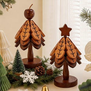 Puede incluir: Dos adornos de árbol de Navidad de madera. Uno está rematado con una manzana, el otro con una estrella. Ambos tienen piezas de madera en forma de hoja. Los árboles son marrones y se asientan sobre bases redondas. Otros adornos festivos los rodean.