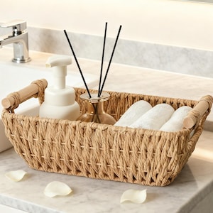 May include: A woven rectangular basket with wooden handles holds a white soap dispenser, a gold-colored diffuser with black reeds, and white washcloths. The basket sits on a marble countertop with scattered white petals.
