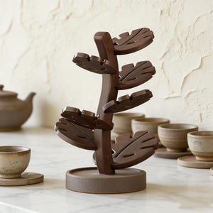 Puede incluir: Un árbol de madera marrón oscuro para tazas y platillos de té. El árbol tiene un tronco central con niveles en forma de hoja para sostener las tazas. La base es de color marrón oscuro a juego. Varias tazas y platillos de té están en el fondo.