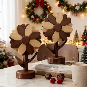 Könnte beinhalten: Zwei Holzblumenskulpturen mit abwechselnd dunkelbraunen und hellbraunen Blütenblättern. Die Skulpturen haben einen Stiel und ein Blattdesign. Der Hintergrund umfasst Weihnachtsdekorationen, Lichterketten und einen Stapel Bücher.