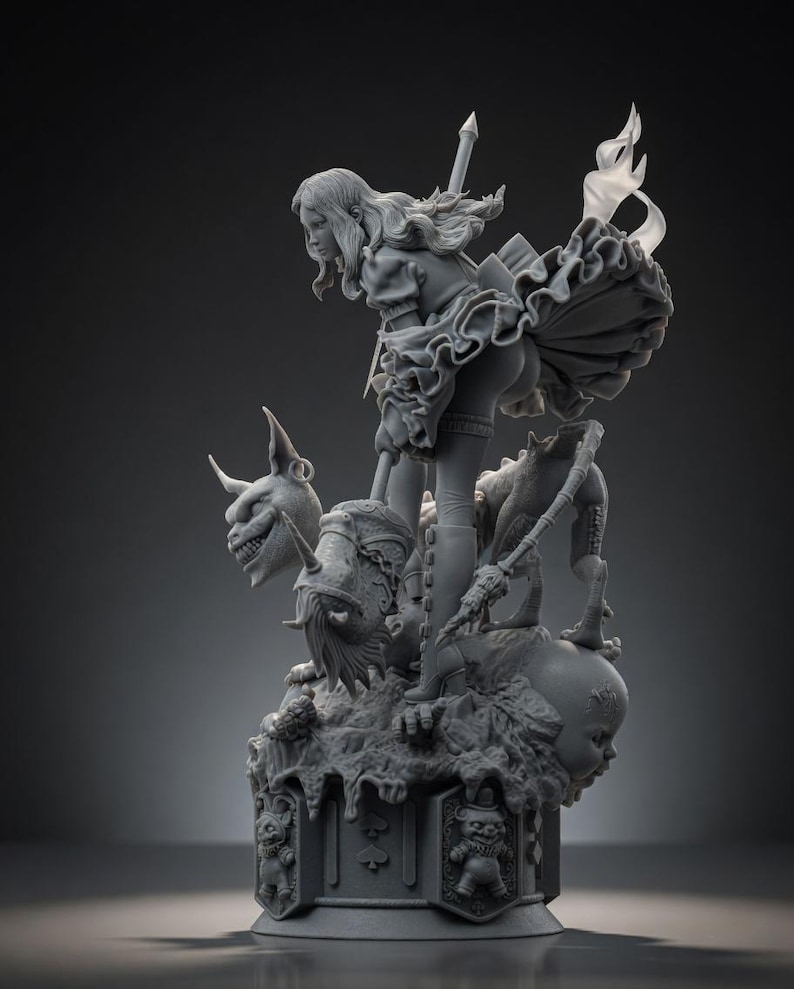 May include: A gray resin figurine depicting a character with long hair holding a long staff, standing atop a base decorated with card suit symbols and small figures. The character wears a ruffled skirt and boots, surrounded by a horned creature and a doll head.