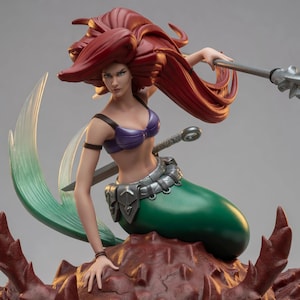 May include: A detailed figurine of a mermaid with long flowing red hair, wearing a purple shell-shaped top. The figure has a green tail, a silver metallic belt, and holds a silver spear. It is positioned atop a textured, dark red rocky base.
