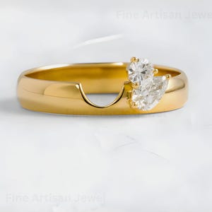 Puede incluir: Un anillo de oro con un diseño único, con dos piedras preciosas claras y brillantes. Una gema es redonda y la otra en forma de pera. El anillo tiene una banda lisa y pulida y un diseño recortado distintivo. El texto "Fine Artisan Jewel" está en la imagen.