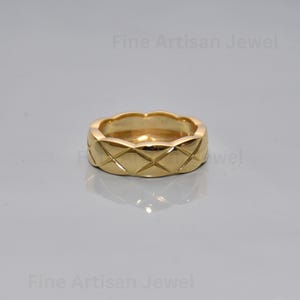 Puede incluir: Un anillo dorado con un patrón acolchado. El anillo tiene una forma redondeada con hendiduras en forma de diamante en su superficie. El texto "Fine Artisan Jewel" es visible. Fondo gris claro.