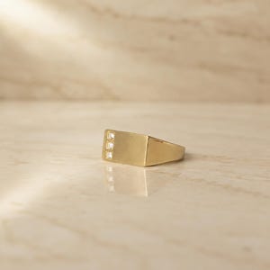 May include: A gold signet ring featuring a flat rectangular top surface with three small, clear, square-cut stones set vertically in a row on the left side. The band has a smooth, polished finish and a solid, tapered construction.