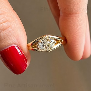 May include: A gold ring featuring a central cushion-cut diamond set within a sleek, polished band. The metal band splits into two distinct arms that frame the gemstone, creating a geometric, modern look with a high-shine, reflective finish.