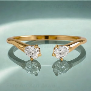 Op de afbeelding: Een goudkleurige ring met twee heldere, peervormige edelstenen. De band van de ring buigt omhoog om elke edelsteen te omsluiten, waardoor een open ontwerp ontstaat. De ring wordt gepresenteerd op een reflecterend oppervlak.