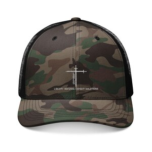 Op de afbeelding: Camouflage trucker pet met een zwarte mesh achterkant en een gebogen klep. Het voorpaneel heeft een wit kruisontwerp met de tekst "LULS" en "LIBERTY DEFENSE COMBAT SOLUTIONS". De pet is een mix van groen, bruin en zwart.