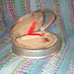 May include: An open, round, silver-toned compact with a clear lid. The interior contains a light-colored powder and a red stripe. The compact sits on a blue fabric with colorful embroidered details.