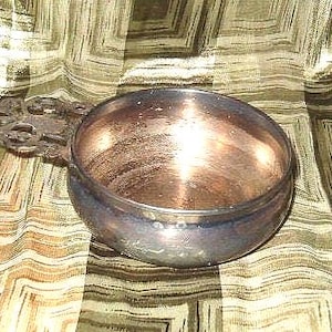 May include: A silver-colored bowl with a decorative handle. The bowl has a rounded shape with a wide opening and a polished interior. The handle features an ornate, openwork design. The bowl sits on a patterned fabric.