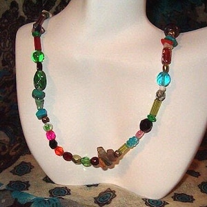 May include: A colorful beaded necklace with a variety of shapes and sizes. The necklace features green, blue, pink, and brown beads, along with other colorful accents. The necklace is displayed on a white mannequin.