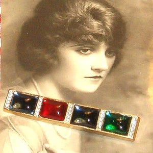 May include: A vintage gold-toned bar pin with four square, colorful stones: blue, red, blue, and green. Each stone is set within a frame of small, clear crystals. The pin is displayed against a sepia-toned portrait of a woman.
