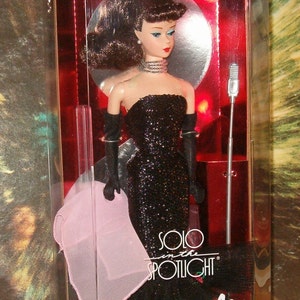 May include: A vintage Barbie doll in a box. The doll is wearing a black sequined gown, long black gloves, and a silver necklace. The box reads "ORIGINAL 1960 FASHION & DOLL! Special Edition Reproduction" and "SOLO in the SPOTLIGHT Barbie."