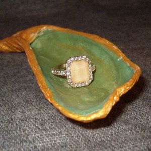 May include: A handmade ring dish in the shape of a leaf, painted green with gold trim. A silver ring with a square, light-colored stone sits in the center of the dish. The ring is surrounded by small, clear stones.