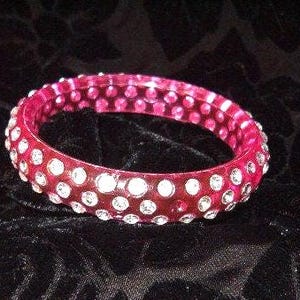 May include: A clear, red bangle bracelet with numerous white, round rhinestones. The bracelet is circular and appears to be made of a transparent material. The rhinestones are evenly spaced around the bracelet.