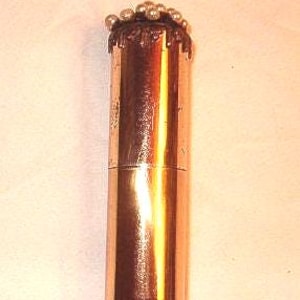 May include: A copper-colored, cylindrical container with a decorative top. The top features a dark, textured band with small, silver-colored spheres clustered around the edge. The container has a smooth, reflective surface.