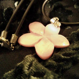 May include: A flower pendant necklace with a light pink, five-petal flower. The pendant is attached to a black cord necklace with a silver clasp. The flower pendant is on a dark green, textured surface.