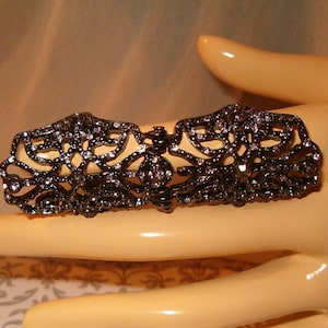 May include: A dark gray filigree ring with clear rhinestones. The ring has an ornate, openwork design with a symmetrical pattern. The ring is a statement piece of jewelry.