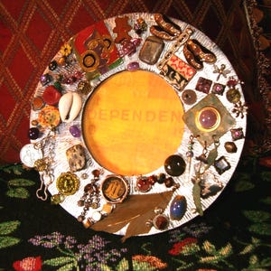 May include: A round, silver-colored picture frame with a yellow center and ornate embellishments. The frame is decorated with various beads, shells, and charms, including a dragonfly and the word "DEPENDEN" in the center.