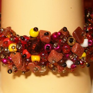 May include: A beaded bracelet with a mix of red, yellow, brown, and white beads and stones. The bracelet is made of various sizes and shapes of beads, including small round beads and larger, square-cut stones. The bracelet is displayed on a cream-colored surface.