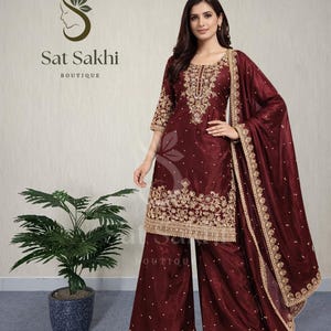 May include: Burgundy-colored palazzo pants suit with gold embroidery. The suit includes a long-sleeved top and a matching scarf. The top and pants are adorned with intricate gold embroidery. The text "Sat Sakhi Boutique" is visible.