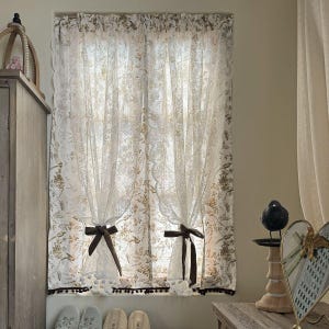 May include: Sheer white window curtains with a floral and bird pattern. The curtains are tied back with dark brown bows and have a scalloped edge. A row of white pom-poms and dark brown trim adorns the bottom.
