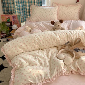 Pode incluir: Um conjunto de cama com capa de edredão e duas fronhas creme com um pequeno padrão floral rosa e bordas com folhos. Um cão de peluche, uma pequena caneca de cerâmica, uma mala de tecido bege e auscultadores brancos estão sobre a cama.