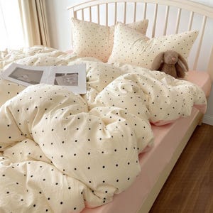 Może przedstawiać: Kremowy zestaw pościeli w czarne grochy, w tym poszwa na kołdrę i poszewki na poduszki, na łóżku z różowym prześcieradłem. Pluszowy królik i czasopismo leżą na łóżku. Rama łóżka jest biała.