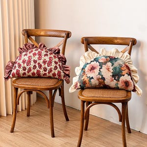 Puede incluir: Dos sillas de madera con asientos tejidos sostienen cada una un cojín decorativo. Un cojín tiene un estampado floral rojo con un borde burdeos con volantes. El otro cojín tiene un estampado floral con flores rosas y blancas y un borde crema con volantes.